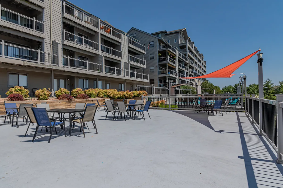 An inviting exterior scene showing tables with colorful umbrellas, flower beds, and the building facade. The setup hints at outdoor dining, social gatherings, and vibrant community events.