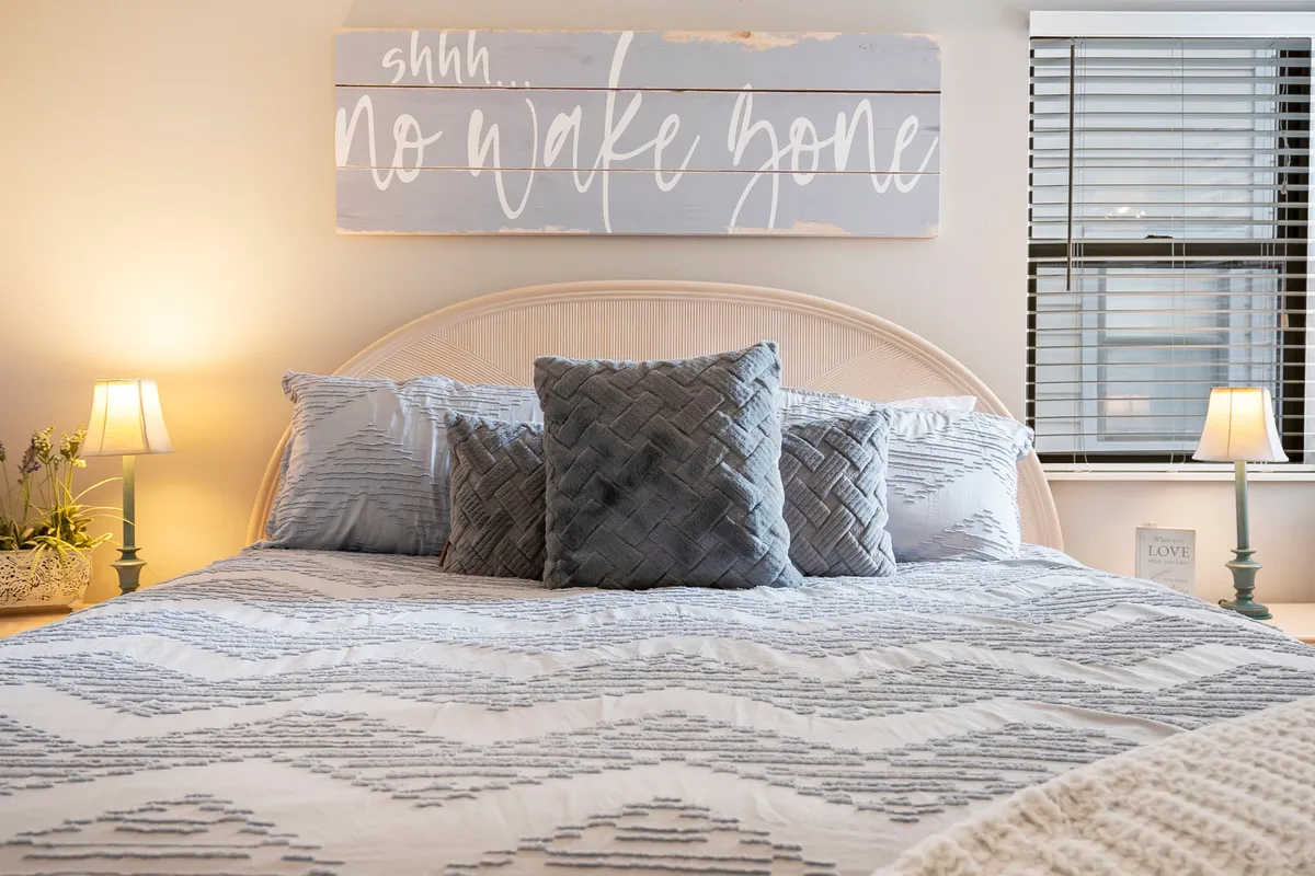 A tranquil bedroom scene with a bed adorned in decorative pillows, a wooden sign above the headboard, and nightstands complete with lamps. The soft lighting and calm ambiance evoke a sense of peace, perfect for a restful night's sleep.