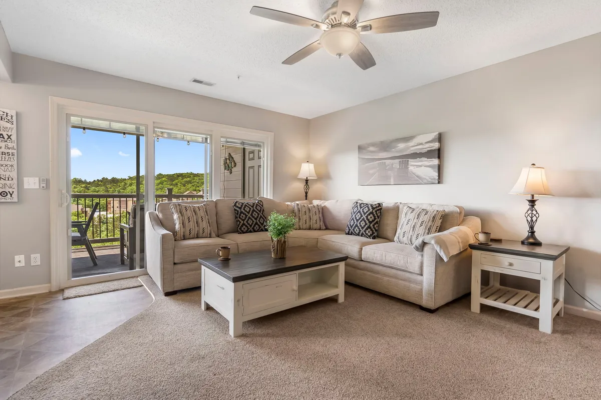 A large sectional sofa anchors a cozy living room, with a coffee table, decorative items, and a lamp creating a warm, homey atmosphere. The image also reveals a view of the balcony, indicating proximity to natural vistas and easy access to outdoor relaxation.