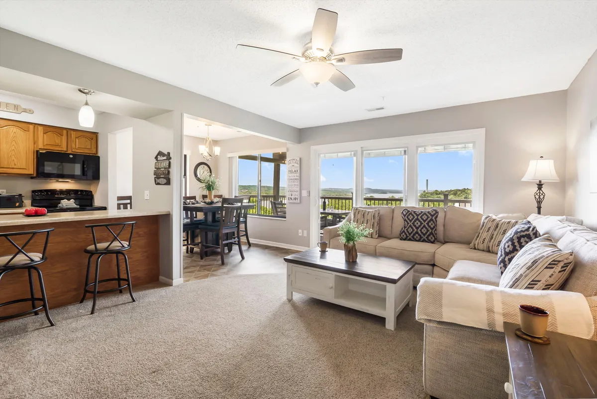 This image features a comfortable open-plan living space with a clear view of both the dining area and kitchen bar. The large windows flood the space with natural light and offer a glimpse of the sparkling lake outside, reinforcing the 'dining with a view' experience. The scene exudes warmth and a welcoming ambiance, perfect for gatherings and shared meals.