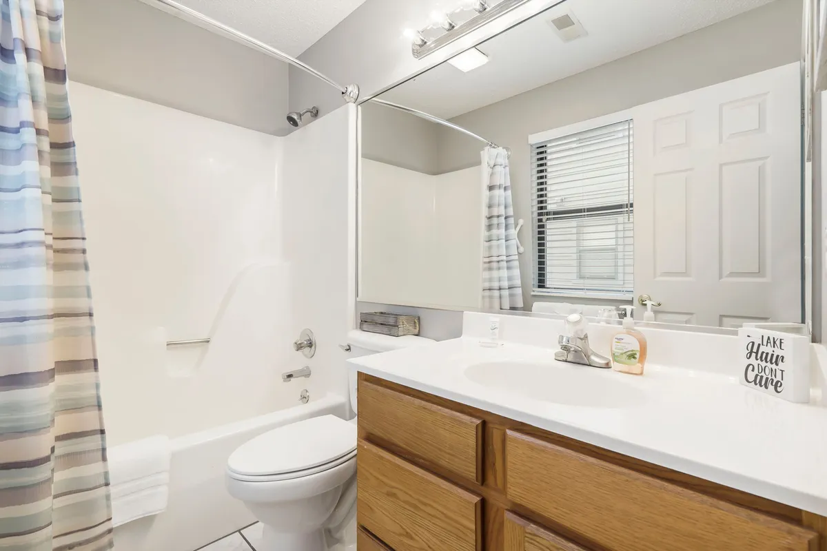 A modern, spa-like bathroom with a vanity, large mirror, bathtub, shower curtain, and a window allowing natural light to brighten the space. The clean lines and serene styling evoke a refreshing, resort-inspired ambiance.