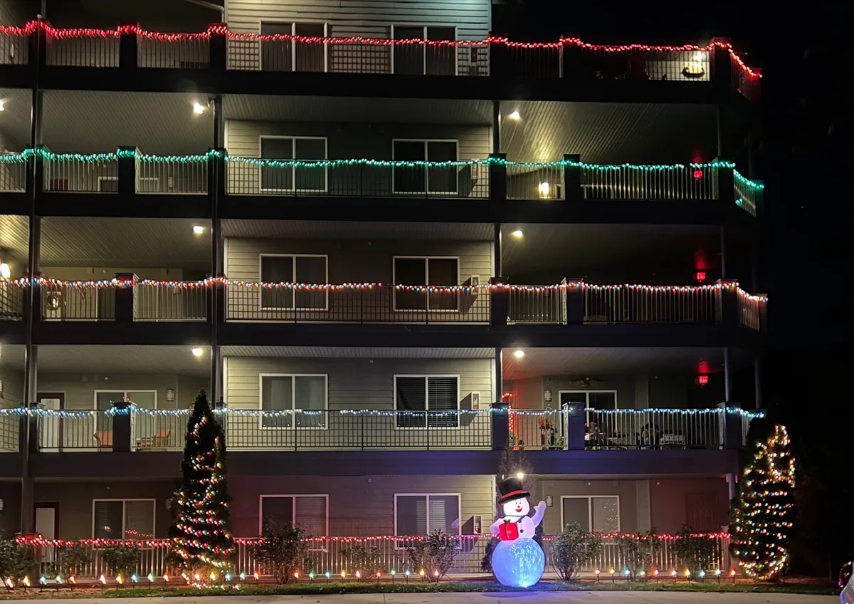 A festive outdoor scene adorned with colorful holiday lights, a snowman decoration, and decorated trees, evoking the joyful spirit of winter celebrations on the property.