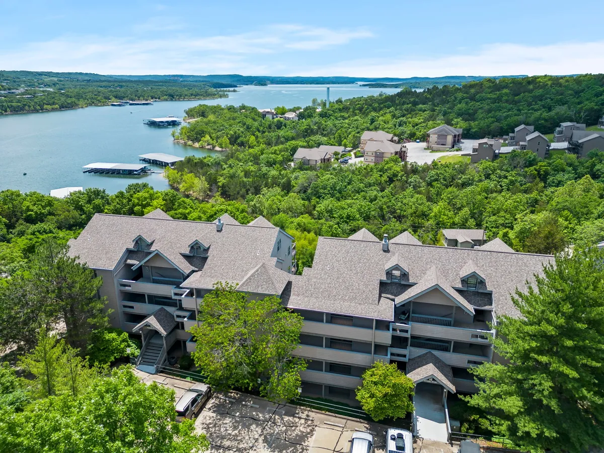 A wide exterior shot featuring the serene lake, lush trees, and the building structure with nearby docks. The scene evokes a peaceful atmosphere perfect for outdoor relaxation, boating, or simply savoring the natural beauty from the property.